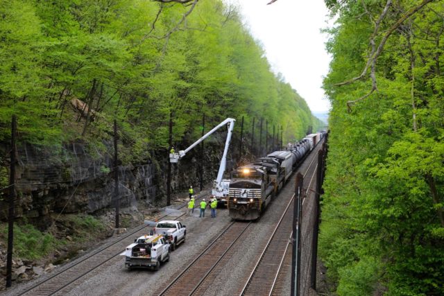 NS eastbound freight creeps past workers fixing a broken slide fence in a deep cut at Cassandra, Pennsylvania. When wires on the fence are broken by rocks or large chunks of ice falling to the track, a red stop signal is automatically displayed to prevent rail accidents. This is the old Pennsylvania Line; the fourth track was where the trucks are positioned. This is about 10 miles west of the Horseshoe Curve.